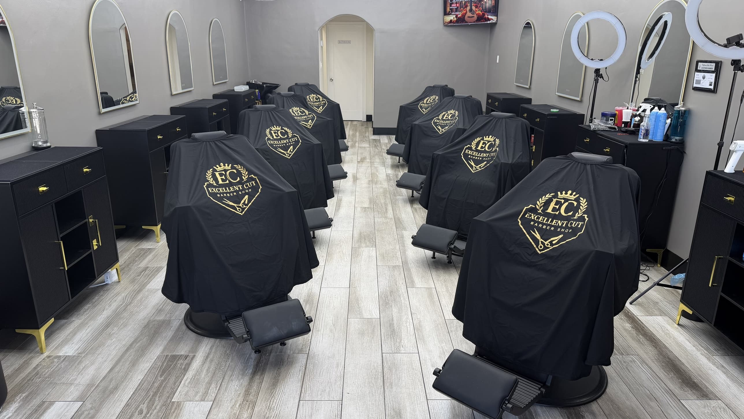 Modern barbershop interior with rows of chairs covered in black capes featuring gold logos.
